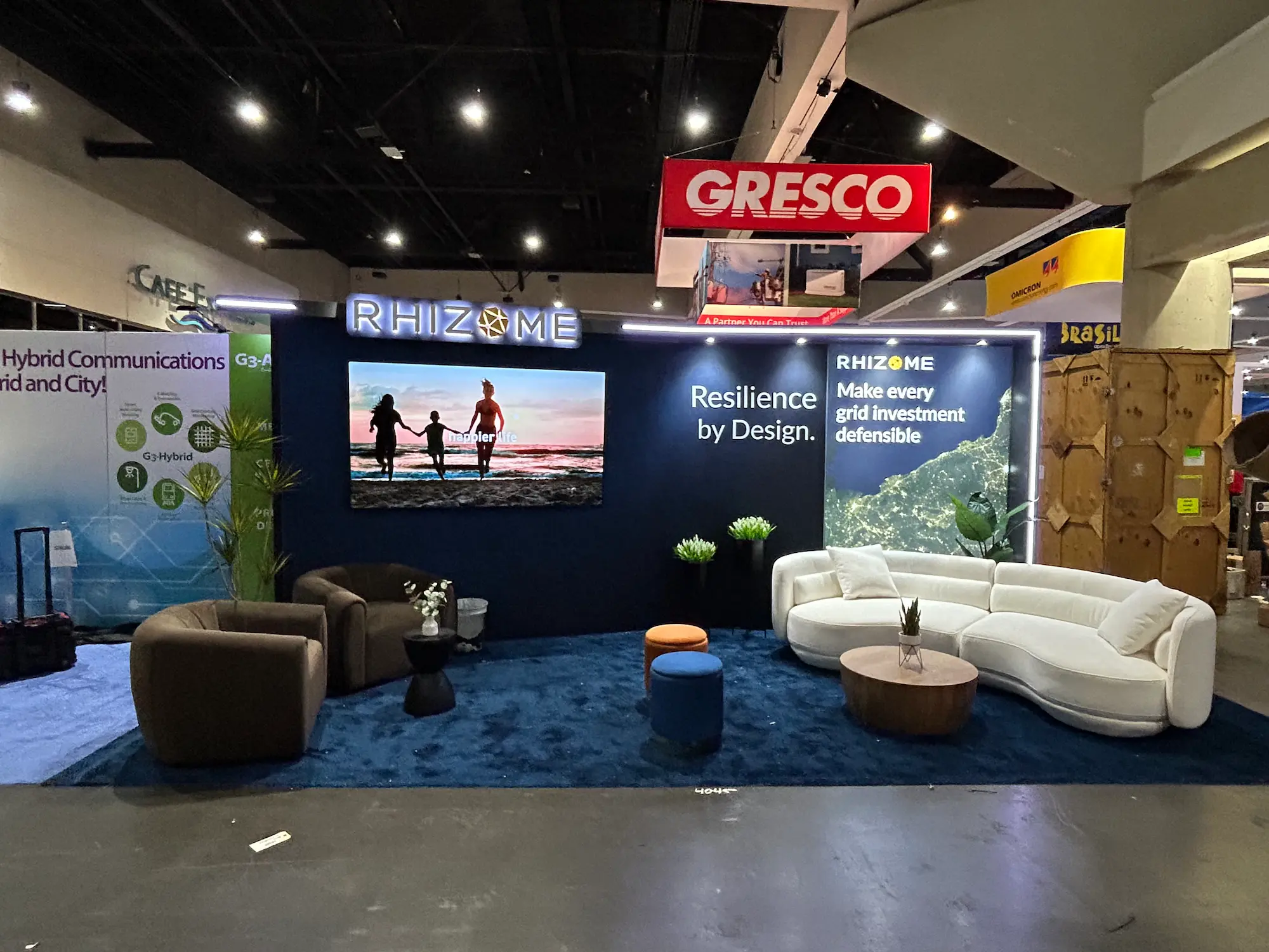 Custom 10x20 trade show booth for Rhizome featuring a navy blue backwall with large LED video display, illuminated brand signage, backlit graphic panels, deep blue carpet, and a furnished lounge seating area with a curved white sofa, accent chairs, and ottomans on the trade show floor.