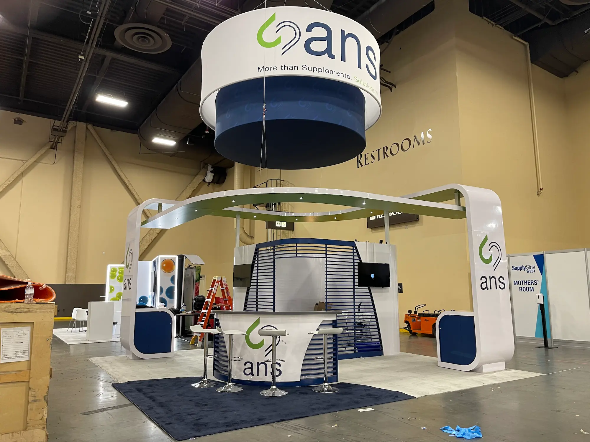 ANS 20x20 island booth design with circular hanging sign, open four-sided layout, and branded counters by Highway 85 Productions